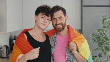 Portrait of handsome mens gay family holding lgbt flag smiling. Attractive romantic male lgbt couple standing in their new home in morning and looking at camera with gay pride and rainbow background