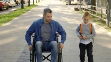 A disabled father in a wheelchair and his son talk about school and how he spent his day after school. The disabled father picked up his son from school and they go home together