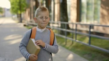Male smiled pupil wearing glasses carrying backpack go to school holding the book. Educated kids. A little schoolboy going to class to learning