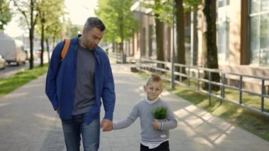 Father carrying backpack holding sons hand and son is holding the plant in a pot after ecology lesson at school smiling wearing glasses. People and ecological concept