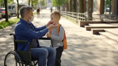 Close up of the disabled father in mask sitting on the wheel chair wearing mask to his son before school because of the pandemic and tells him the rules. Pandemic and disabled people concept