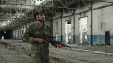 Strong military soldier with big rifle in his hands walking through abandoned old building. Member of armed forces guarding and patrolling destroyed steel factory.