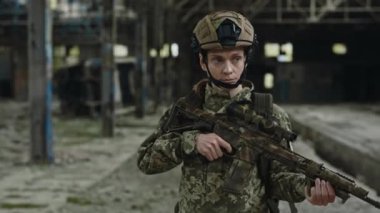 Professionally trained female soldier walking at destroyed plant and using military rifle for looking in sight. Armed caucasian woman wearing uniform and helmet.