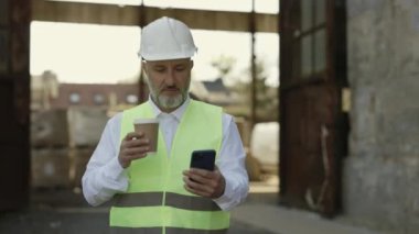 Kafkasyalı olgun ustabaşı modern akıllı telefonlarda internet taraması yapıyor ve inşaat alanında yürürken sıcak kahve içiyor. Miğferli sakallı mimar ve yansıtıcı yelek iş yerinde mola veriyor..