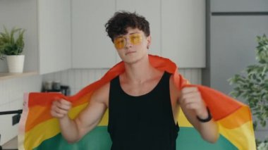 Portrait shot of young stylish gay man wearing yellow glasses and rainbow flag over his shoulder and looking at camera while posing. People and lgbt concept