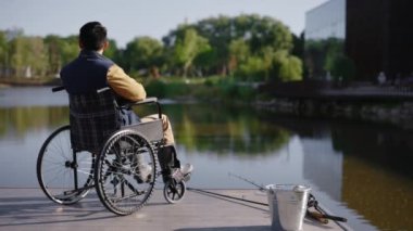Back view of the disabled fisherman sitting on the wheelchair in the pier looking at the local lake after fishing alone. Disabled people and fishing concept