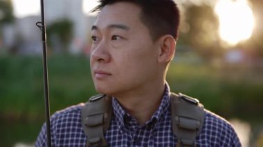 Close up of the smiled fisherman wearing special clothes for fishing holding the fishing rod standing looking at the camera on the blurred lake background. People and leisure time, fishing concept