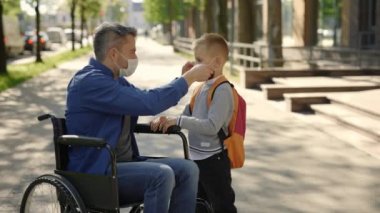 Disabled father in mask sitting on the wheel chair wearing mask to his son before school because of the pandemic and son goes to the school. Pandemic and disabled people concept