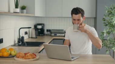 Handsome man is in his new home sitting on the chair in kitchen using laptop for distance work and drinking coffee while working. Technology and people concept
