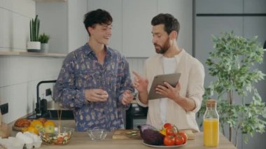 Lgbt couple in love standing together in their new apartment in the kitchen and cooking, one partner showing different receipts using the tablet and another breaks eggs. Family cooking