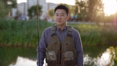 Portrait of the smiled fisherman wearing special clothes for fishing holding the fishing rod standing looking at the camera on the blurred lake background. People and leisure time, fishing concept