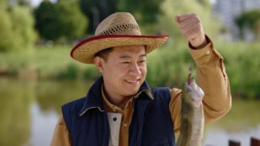 Close view of the asian man wearing special fishing clothes holding the fish he caught on the hook and feeling happy about it looking at the camera