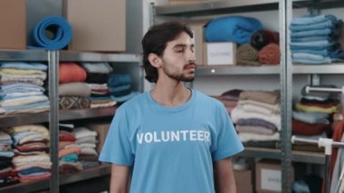 Portrait view of the caucasian man in volunteer t shirt looking at the camera with smile. Shelves with belongings for donating at the background. Humanitarian aid concept