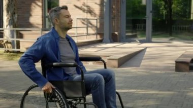 Side view of the happy disabled man in wheelchair is riding in city alone moving wheels with hands in the middle of the day. Handicapped people and outdoor activity concept