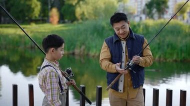 Asian dad shows his son how to fish properly, gives instructions about fishing. Family spend time actively, father tells his kid how regulate the fishing rod