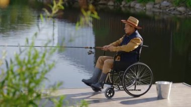Side view of the disabled asian man wearing hat and special fishing clothes sitting on the wheelchair fishing from the pier on the local lake. Disabled people and fishing concept