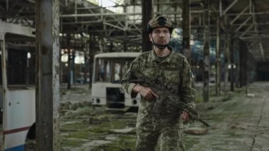 Serious military man in uniform with machine gun patrolling inside broken building. Strong army soldier looking around while walking through collapsed factory.