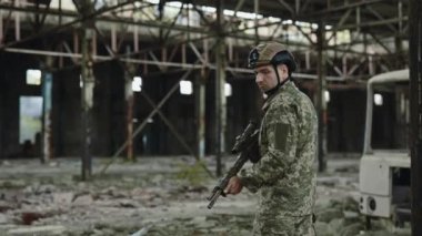 Portrait of caucasian soldier in protective helmet holding precision weapon and looking at camera with calm face. Devoted man executing military mission in abandoned manufacture place