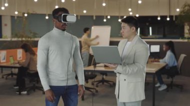 It company. African american man testing a VR simulator headset and while his male colleague data analysis and recording on a laptop. People working at office. Data science