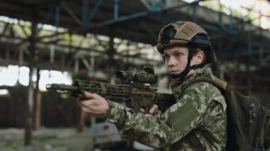 Caucasian woman in camouflage clothes and helmet looking through sight of rifle while standing on damaged manufacture plant. Military training, preparation for war.