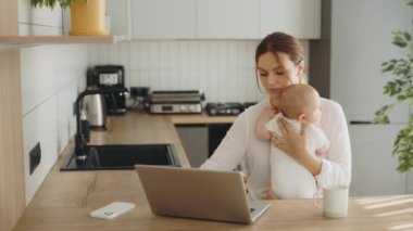 Business worried mother working at the computer holding a newborn baby on her lap sitting in the kitchen at the table. Technology, working and motherhood concept. Home office