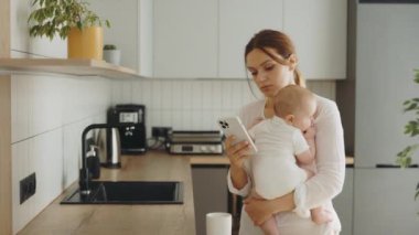 Anxious mother using smartphone the phone looking back at the baby she is holding in her arms. Caucasian businesswoman is texting on the phone while holding her child in her arms