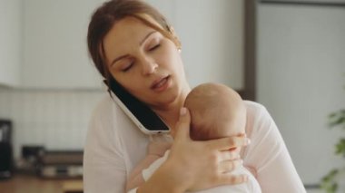 Busy mother talks on the smartphone, solves important matters and holds a newborn baby in her arms. Young business woman preoccupied with work on the phone at work. Home office