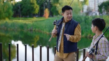 Asian father tells his son how to fish properly, gives instructions about fishing. Father and son spend time actively, dad tells his son about fishing. Family and father-son relationship concept
