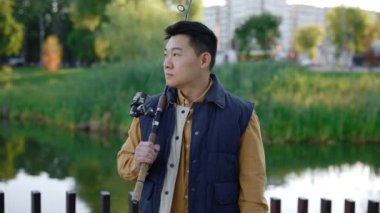 Smiled asian fisherman wearing waistcoat holding the fishing rod standing looking at the camera on the blurred local lake background. People and leisure time, fishing concept