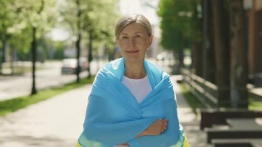 Mature woman carries a the ukrainian flag standing on the street looking at the camera on the blurred background. People portrait. Political concept