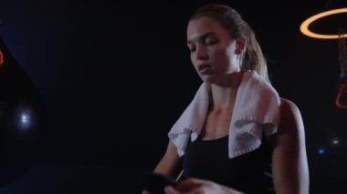 Portrait of the boxer woman binds hand bandage before the training standing in the boxing gym carrying the white towel on her shoulders looking at the camera. People and sport concept