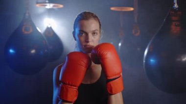 Close up of young attractive female boxer in gloves standing in striking pose and looking at camera at dark ring. Portrait of woman kickboxer ready to punch. Boxing concept