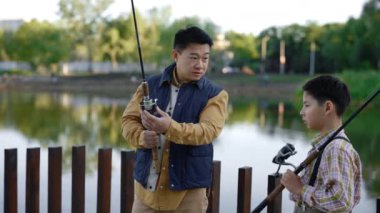 Asian father shows his son how to fish properly, gives instructions about fishing. Father and son spend time actively, dad tells his son how regulate the fishing rod