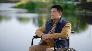 Serious asian disabled man sitting on the wheelchair on the pier in the local lake looking at the camera. People and rehabilitation concept