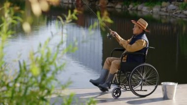 Side view of the disabled asian man wearing hat and special fishing clothes sitting on the wheelchair and cast a fishing rod. Disabled people and fishing concept