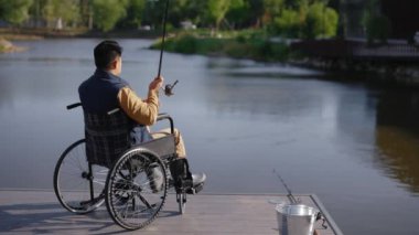 Back view of the male fisherman fishing on the pier early in the morning in the local lake alone. Disabled people and fishing concept