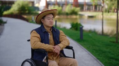 Close up of the serious asian disabled man sitting on the wheelchair on the street near the local lake looking at the camera. Disabled people and rehabilitation concept