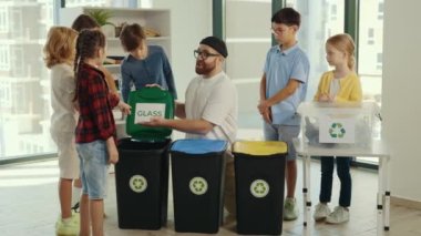 Çöp sınıfında, genç bir öğretmen çocuklara çöpleri nasıl ve nerede ayıklayacaklarını anlatıyor, sınıflandırmayı kolaylaştırmak için yazıtlar gösteriyor ve çocuklara parmaklarını gösteriyor.