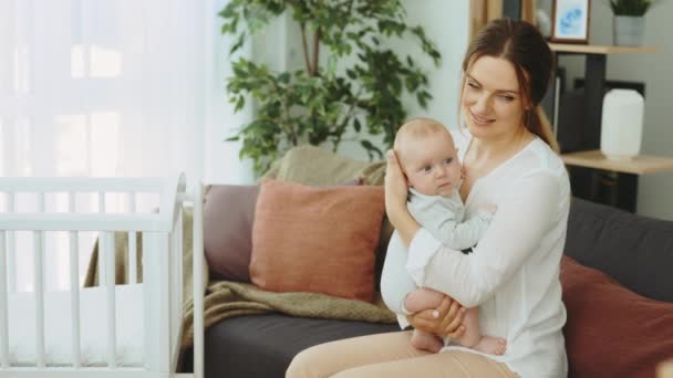 照片上的母亲微笑着坐在新家沙发上抱着新生儿 母亲抱着小儿子坐在靠近婴儿摇篮的沙发上 — 图库视频影像