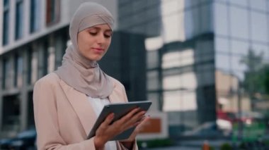 Kahverengi tesettürlü mutlu Arap iş kadını dışarıda çalışıyor, sosyal medyayı araklıyordu. Genç Arap bayan iş merkezinin arka planında tablet üzerinde çalışıyor.