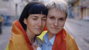 lgbt bayrağına sarılmış kameraya bakan gülümseyen lezbiyen bir çiftin portresi. Şehir merkezinde gökkuşağı bayrağıyla poz veren iki mutlu bayan arkadaş. Kapatın. Kadın ilişkileri, lgbt kavramı