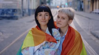 Igbt bayrağına sarılmış lezbiyen bir çiftin portresi şehir merkezindeki caddede duran kameraya bakıyor. Gökkuşağı bayrağıyla poz veren iki mutlu bayan arkadaş. Kadın ilişkileri, Igbt konsepti.