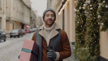 Beyaz bir adam elinde kız arkadaşı için elinde kahve ve noel hediyesi olan gri şapkalı ve eşarplı bir şekilde sokağa bakıyor. İnsanlar ve Noel tatili konsepti