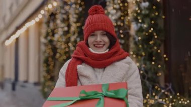 Kırmızı şapkalı mutlu beyaz kadının portresi süslenmiş sokakta elinde büyük bir Noel hediyesiyle dikilmiş eşarba sarılmış. Kapat. İnsanlar ve Noel tatili konsepti.