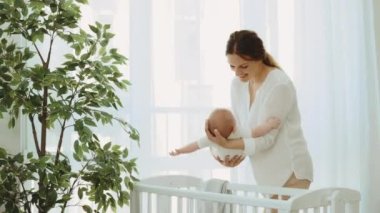 Neşeli beyaz anne, yeni doğmuş bebeğini elinde sallar. Pencere arkasında bebekle oynar. Annelik kavramı