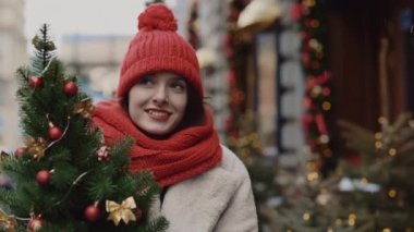 Mutlu gülümseyen kadın kırmızı şapkalı ve atkıyla süslenmiş arka planda küçük bir ağacı taşıyan Noel yürüyüşünden zevk alıyor. Kapat.