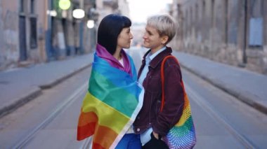 Sokakta dikilmiş kameraya bakan Igbt çifti portresi. İki lezbiyen kadın kameraya bakıyor. Kadın ortak, kız arkadaşının yanında lgbt bayrağını taşıyor.