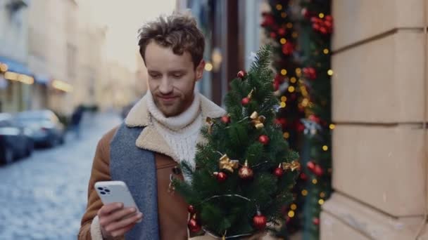 スマートフォンを使ってクリスマスツリーの小さなクリスマスツリーを持ち アプリを交換し ガールフレンドとテキストメッセージを送るクリスマスツリーショップから帰宅している笑顔の白人男性 人と技術の概念 — ストック動画