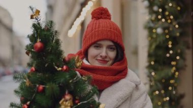 Atkıya sarılmış kırmızı şapkalı güzel beyaz kadının portresi bulanık caddedeki kameraya bakıyor. Kapatın. İnsanlar ve Noel tatili konsepti