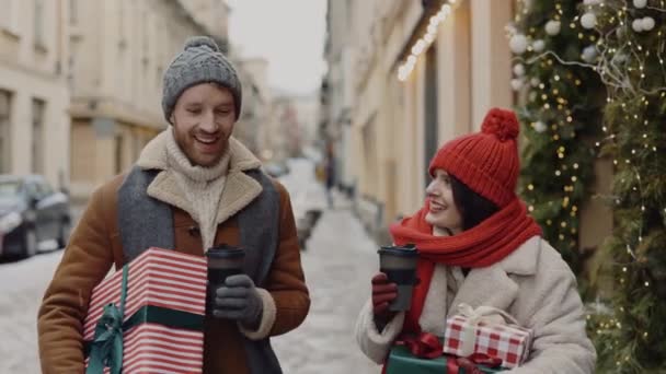 白人のカップルが歩いている 素敵な話すと一緒にクリスマスの贈り物を運ぶ 女性はコーヒーを飲んでいます 白人のカップルはクリスマスの買い物から家に帰る予定です — ストック動画